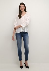 Blusa bianca con dettagli in pizzo sulle maniche, scollatura a V, abbinata a jeans skinny blu e scarpe con tacco nere, su uno sfondo neutro.