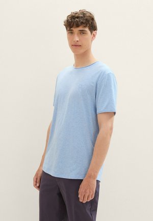 Lyseblå kortermet T-skjorte i et mykt materiale med rund hals og en liten logo på brystet. Modellen har på seg mørke bukser.