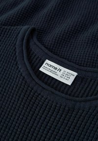 Granatowy sweter z prążkowanej dzianiny o okrągłym dekolcie. Metka pokazuje rozmiar i instrukcje pielęgnacji, widoczne są detale struktury materiału.