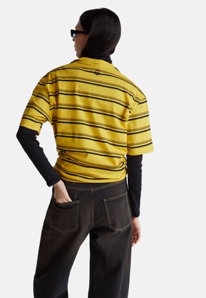 Camicia a maniche corte con righe gialle e nere, abbinata a pantaloni di jeans scuri con due tasche posteriori e dettagli di cuciture visibili.