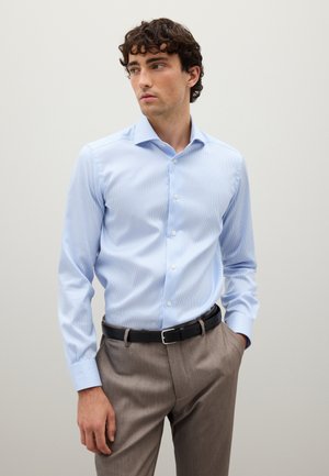 Camisa de vestir a rayas verticales azul claro, con cuello clásico y mangas largas, combinada con pantalones color arena y un cinturón negro.