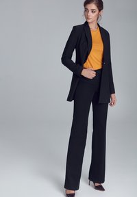 Kobieta ubrana w czarną marynarkę, czarne spodnie, musztardowy top i czółenka z ostrym noskiem, stojąca na tle jasnoszarym.