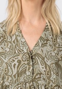 Vaalea nainen, joka pitää ykkösvartalon mekkoa, jossa on vihreä, beige ja valkoinen paisley-kuvio, paljastaen niskan ja ylärinnan.