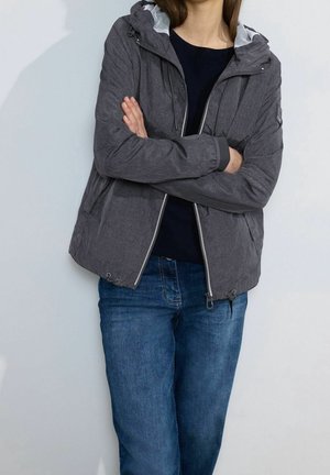 Person iført en mørkegrå hættetrøje over en sort skjorte og blå jeans, med armene krydset foran en ensartet lys baggrund.