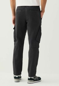 Black cargo pants made of cotton blend, featuring two large side pockets, a relaxed fit, and cuffed hems. Worn with black and green sneakers.