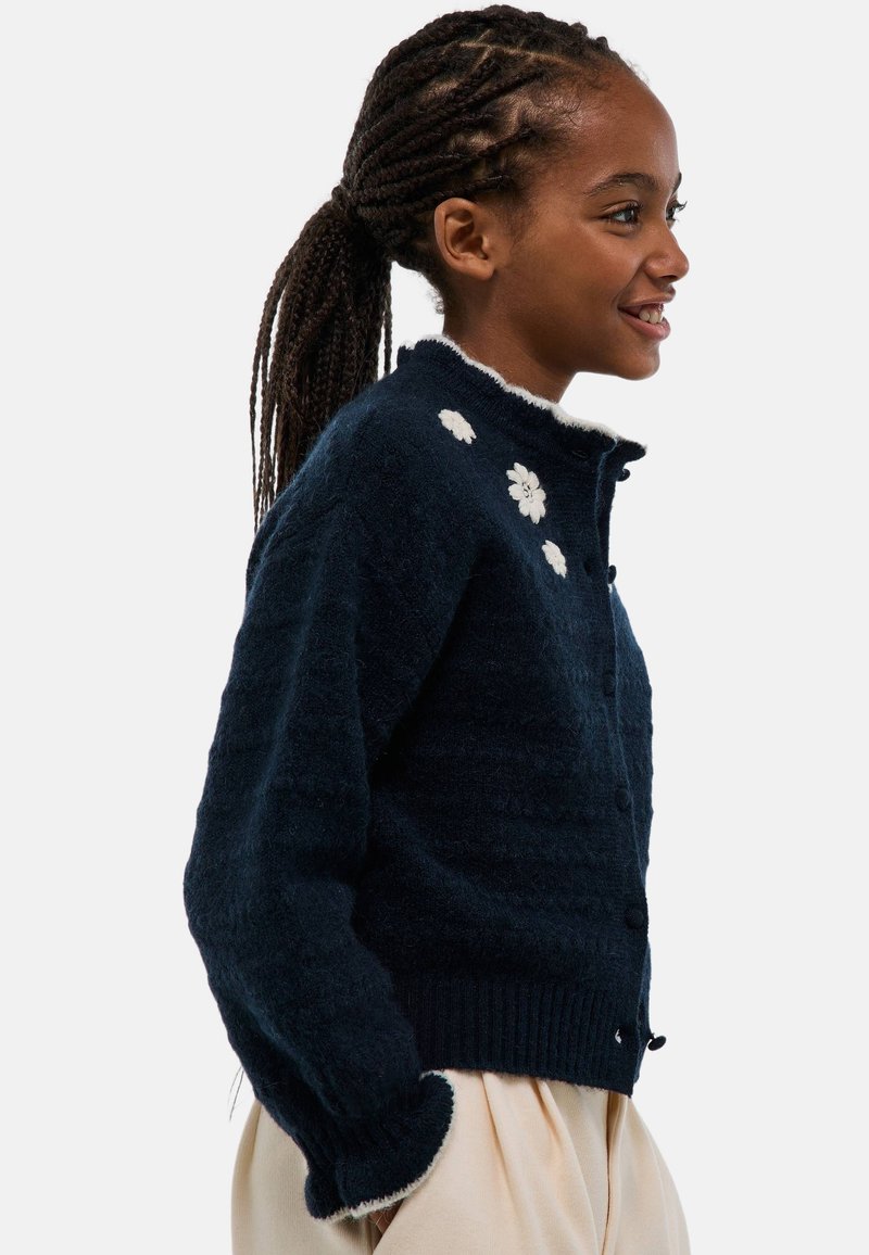 Cardigan en maille marine avec une finition texturée, orné de broderies de fleurs blanches, avec un col rond et des poignets et un ourlet côtelés.