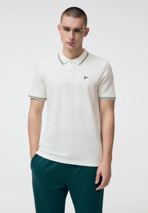 Młody mężczyzna ubrany w biały polo z krótkim rękawem i paskowanymi brzegami oraz ciemnozielone spodnie w wzory, stojący na jednolitym tle.