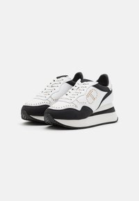 Baskets compensées blanches et noires avec lacets, ornements en forme de clous dorés formant un dessin circulaire sur les côtés, présentées sur un fond uni.