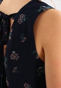 Marinebluse mit Blumenmuster und Bindeband am Hals, ausgestattet mit kleinen rosa und grünen Blumendesigns. Weicher, fließender Stoff mit einer glatten Textur.