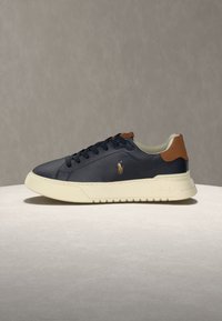 Baskets en cuir noir avec semelle blanche, languette de talon marron et petit logo brodé sur le côté, présentées sur une surface ronde et claire.