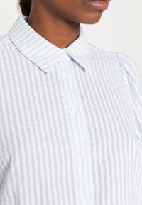 Chemise à rayures bleu clair et blanches avec col classique et tissu texturé. Dotée de boutons blancs et de manches bouffantes.