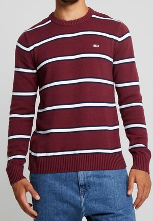 Uomo che indossa un maglione bordeaux a coste con righe orizzontali bianche e blu navy e jeans blu, mostrato dal collo ai fianchi.
