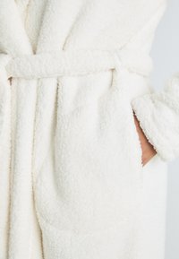 Weißer, flauschiger Bademantel aus einem strukturierten, samtigen Stoff. Mit einem Gürtel in der Taille und seitlichen Taschen sowie aufgerollten Manschetten.