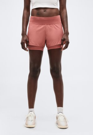 Osoba oblečená v vrstvených atletických šortkách svetloružovej farby, bielom crop top tričku a bielych teniskách stojaca pred jednoliatym pozadím.