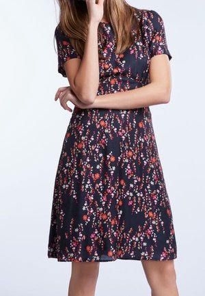 Mujer con un vestido oscuro de manga corta y largo hasta la rodilla, con un patrón floral en tonos rojos, rosas y blancos, de pie con un brazo cruzado.