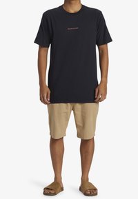 Marinblå bomulls t-shirt med korta ärmar och rund halsringning, med en liten orange logotyp på bröstet. Kombinerad med beige shorts och bruna sandaler.