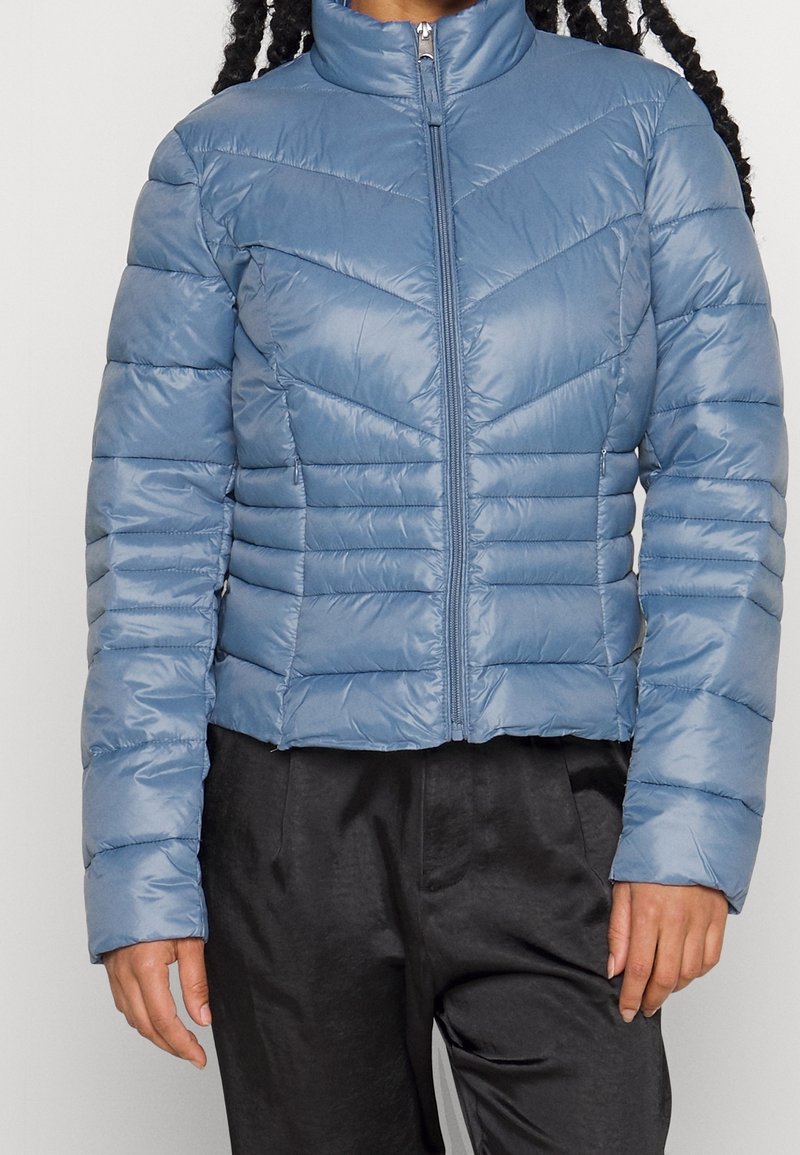 Ljusblå kort pufferjacka med hög krage, chevronmönstrad sömnad, dragkedjestängning och elastiska muddarna. Slät, glänsande yta.