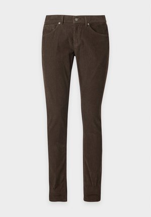 Junger Mann, der mit einer Hand in der Tasche steht und ein schwarzes kurzärmeliges Polo, dunkelbraune Hosen mit aufgekrempelten Bündchen, weiße Socken und schwarze Schuhe trägt.
