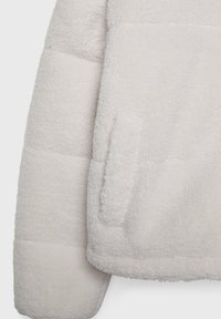 Gros plan sur la manche blanche texturée d'une veste polaire et la poche avant, montrant un matériau épais, doux et matelassé, adapté aux temps froids.