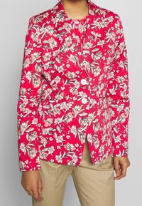 Blazer floreale in un vivace rosa con motivi crema, pesca e verde. Presenta un colletto a punta, due bottoni e tasche frontali. Vestibilità sartoriale.