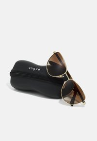Aviator-aurinkolasit ruskeilla linsseillä ja kultaisella metallikehyksellä lepäävät mustassa, vetoketjullisessa kotelossa, jossa on valkoisella kohokuvioitu teksti "vogue".