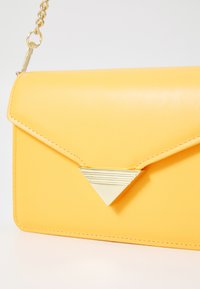 Gul faux skinn clutch-veske med en triangelformet gulldetalj, som har klafflås, sydde kanter og en kjede-rem.
