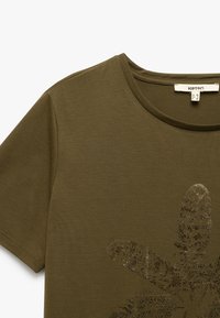 Camiseta de algodón verde oliva con cuello redondo, mangas cortas y un patrón de hojas texturizado con detalle metálico en la parte delantera.