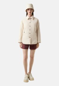 Beżowa pikowana kurtka z kołnierzykiem, zapinana na guziki z przodu, z bocznymi kieszeniami, noszona z bordowymi szortami i beżowymi butami na platformie.