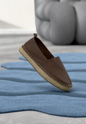 Chaussure espadrille marron à enfiler avec une semelle en jute tressée flottant au-dessus d'un tapis bleu texturé à l'intérieur avec du mobilier gris flou en arrière-plan.
