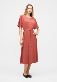 Γυναίκα με κυματιστά μαλλιά, ντυμένη με μίντι φόρεμα σε σκουριά κόκκινο χρώμα, με κοντά μανίκια και ζώνη στη μέση, στέκεται με το ένα χέρι στη μέση και φοράει μαύρες μπαλαρίνες.