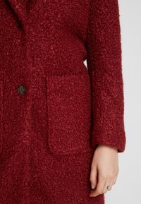 Abrigo rojo texturizado con una superficie suave y peluda; cuenta con dos bolsillos delanteros, cierre de botones y un ajuste relajado.
