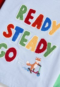 Erinevates tekstuurides värvilised tähestikud "READY STEADY GO" helesinisel kangal, millel on broderitud rebane, kes sõidab punasel ja rohelisel rolleril.