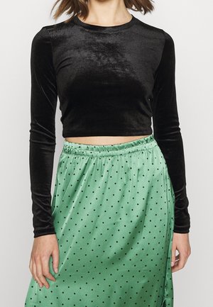Femme portant un crop top en velours noir à manches longues et une jupe en satin verte taille haute à pois noirs.
