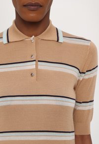 Polo shirt beige à manches courtes avec col, présentant des rayures horizontales en bleu, blanc et marine. Patte de boutonnage à trois boutons et texture douce.