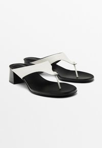 Schwarze Sandalen mit hohen Absätzen und weißen Riemen, die einen t-förmigen Zehensteg und einen klobigen Absatz haben. Das Obermaterial ist glatt und steht im Kontrast zur matten Innensohle.