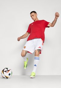 Raudona Adidas marškinėliai, balti šortai su „Manchester United“ logotipu, akcentuoti oranžine spalva. Geltoni sportbačiai, dryžuotos mėlynos ir baltos kojinės, netoliese futbolo kamuolys.