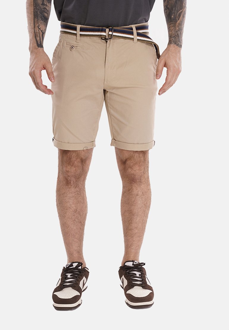 Pantaloni corti beige con risvolti, dotati di chiusura frontale con bottone e zip. Abbinati a una cintura a righe e scarpe da ginnastica marroni.