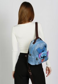 Mochila de tela azul con un diseño de personaje de dibujos animados, que presenta un bolsillo delantero con cremallera, correas marrones y una forma redondeada en general.