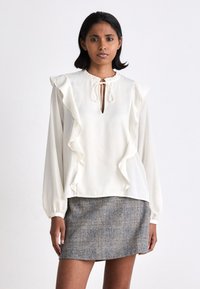 Balta blūze ar ruffle detaļām, atslēgas cauruma dekoltē un garām piedurknēm, apvienota ar pelēku rūtainu svārku. Gluda auduma, ikdienišķs dizains.