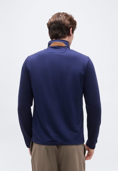 Άνδρας που φοράει ναυτικό μπλε μακρυμάνικο πουλόβερ με καφέ μπαλώματα στον πίσω γιακά, γυρισμένος με το πρόσωπο προς τα πίσω, στέκεται μπροστά σε ένα απλό φωτεινό φόντο.