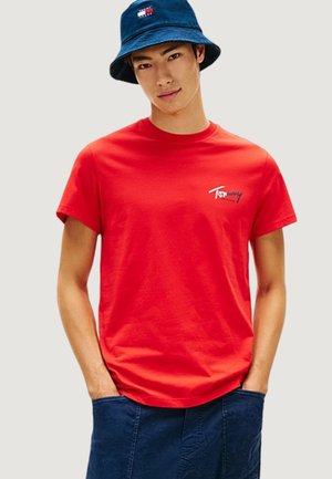 Giovane uomo che indossa una T-shirt rossa Tommy Jeans, un cappello a secchiello blu navy con logo e pantaloni blu navy con le mani nelle tasche.