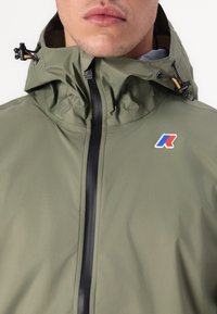 Λαδί αδιάβροχο μπουφάν με ψηλό γιακά, μαύρο φερμουάρ και λογότυπο K-Way. Διαθέτει ρυθμιζόμενα κορδόνια και λεία υφή.