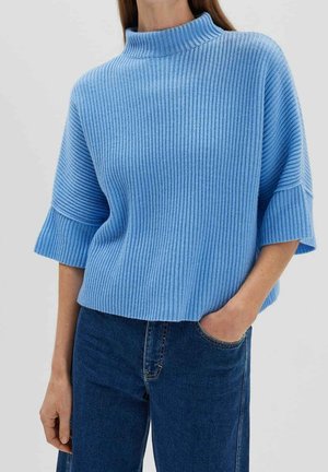 Kvinde iført en lyseblå ribstrikket kortærmet sweater med høj hals og mørkeblå jeans, den ene hånd i lommen.