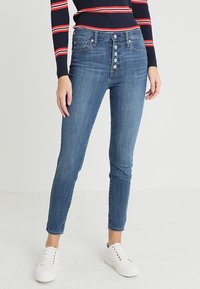 Smal passform blå denimjeans med hög midja, fem-knappsförslutning och subtil blekning. Matcher med vita sneakers för kontrast.