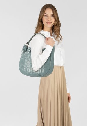 Borsa turchese lucida con texture a coccodrillo, dalla forma curva e con un singolo manico a tracolla, tenuta da una modella in camicia bianca e gonna beige.