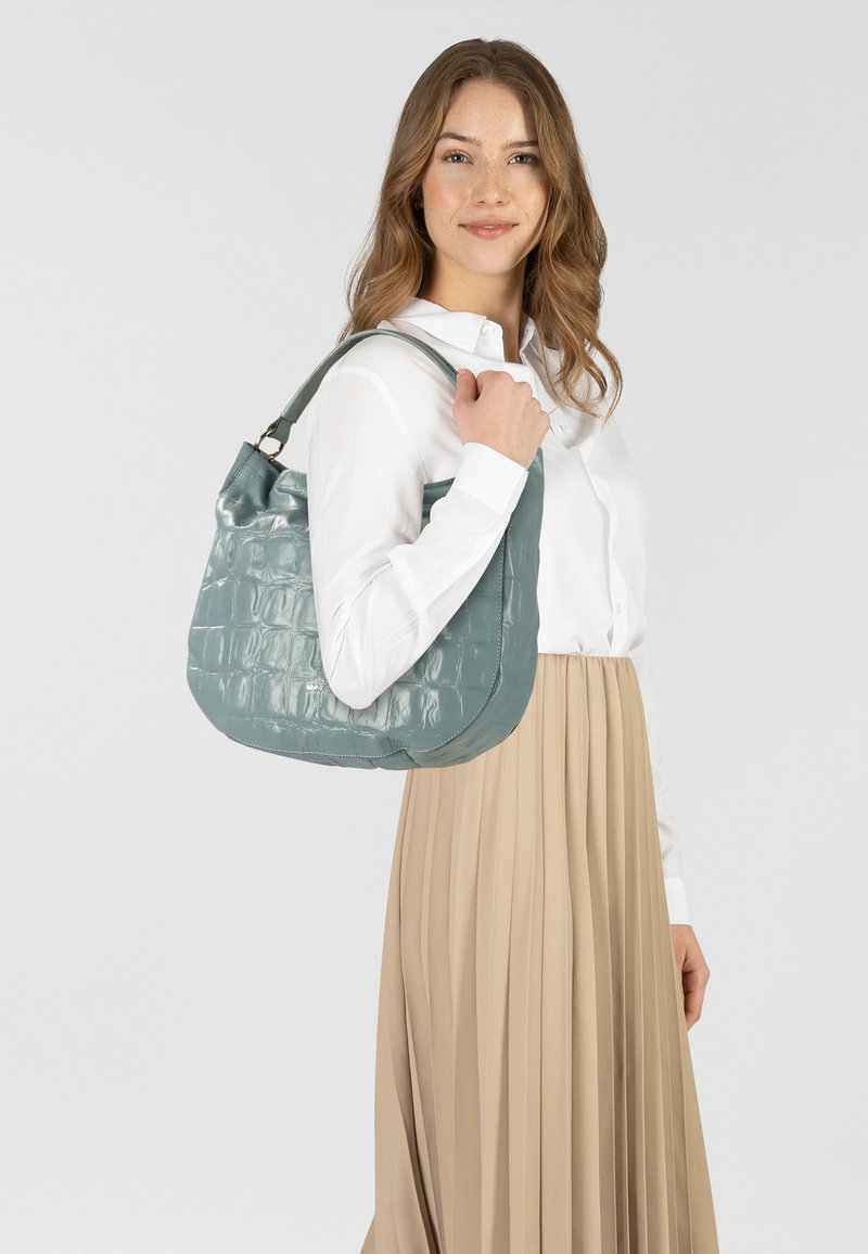 Borsa turchese lucida con texture a coccodrillo, dalla forma curva e con un singolo manico a tracolla, tenuta da una modella in camicia bianca e gonna beige.