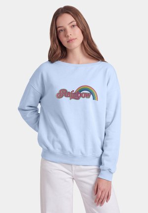 Giovane donna con lunghi capelli castani indossa una felpa azzurro chiaro con arcobaleno colorato e testo "Rainbow", abbinata a pantaloni bianchi, in piedi davanti a uno sfondo bianco.