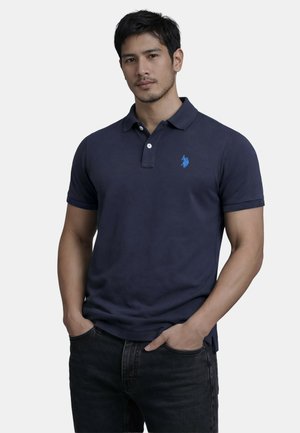 Mann trägt ein marineblaues Polo-Shirt mit einem kleinen blauen gestickten Logo auf der Brust und schwarzen Jeans, Hände in den Taschen, neutraler Hintergrund.