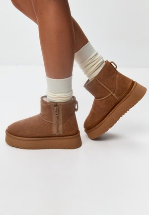 Botine din suede maron cu talpă plată, fermoar pe lateral și cusături texturate. Asortate cu șosete albe cu model în dungi.