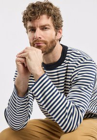 Gestreiftes langärmeliges Hemd in Marineblau und Creme, mit einem runden Kragen in Marineblau. Das Modell hat lockige Haare und einen Bart und sitzt mit gefalteten Händen.